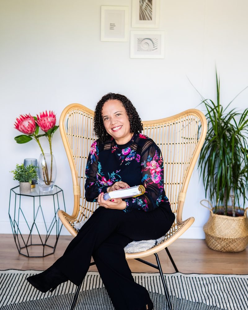 Alicia Noble seated in a wicker chair, holding a book